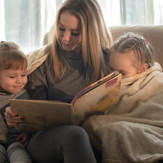 En kvinde sidder i en sofa med to børn. De deler et tæppe og læser i en bog sammen.