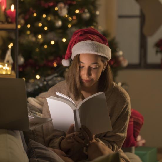 Kvinde, der sidder foran sin computer og læser i en bog. Hun har nissehue på, og der er et juletræ i baggrunden.