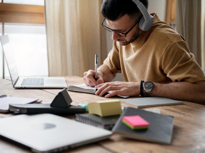 Mand, der skriver på en notesblog. Der ligger papirer, post-its og en computer rundt på skrivebordet.