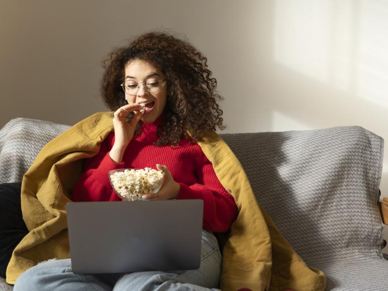 Kvinde, der sidder i en sofa med tæppe på. Hun ser film på sin laptop og spiser popcorn.